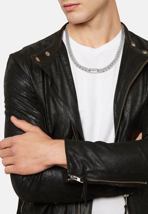 Hombre que lleva un collar de cadena de plata con etiqueta "BREIL", chaqueta de cuero negra con detalles de cremallera y camiseta blanca, con los brazos cruzados.