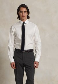 Valge särk terava kraega, mille juurde on sobitatud õhuke, tekstuuriga must lips. Hallid püksid ja pruun vöö, mudel seisab neutraalses taustas.