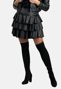 Czarna spódnica z ekoskóry z warstwowymi falbanami, połączona z czarnymi skarpetkami do kolan i botkami na obcasie. Spódnica ma dopasowany pasek w talii.