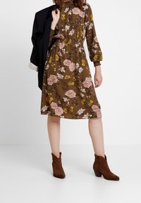 Braunes Kleid mit Blumenmuster, langen Ärmeln, elastischer Taille und knielangem Saum. Pinke und gelbe Blumenmuster auf strukturiertem Stoff. Braune Stiefeletten.