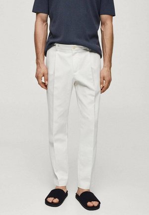 Homme portant un pantalon blanc slim, une chemise bleu marine à manches courtes et des sandales noires ouvertes, debout devant un fond clair uni.