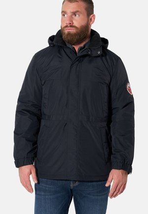 Mann mit Bart, der eine schwarze Winterjacke mit Kapuze und einem Aufnäher am Ärmel sowie blaue Jeans trägt und nach links schaut.