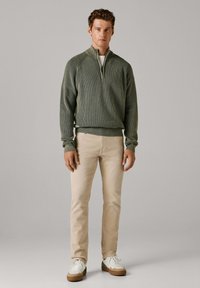 Khakifarbener, gerippter Pullover mit halbhohem Reißverschlusskragen, kombiniert mit beigen Hosen und weißen Turnschuhen mit braunen Akzenten.