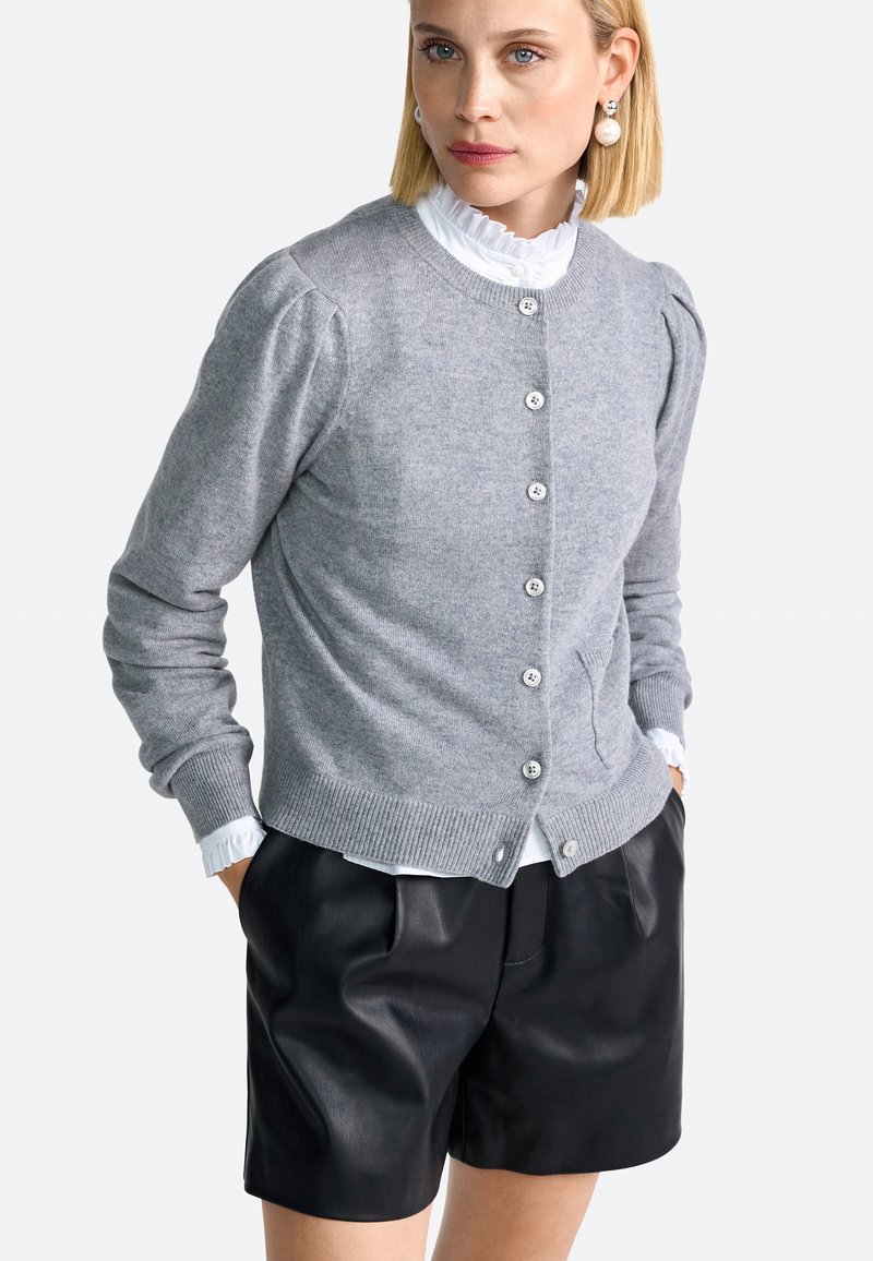 Vrouw in een grijze knopentrui over een witte gerimpelde blouse, met zwarte leren short en parel hangers.