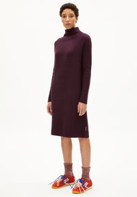 Robe en turtleneck côtelée de couleur violet profond, avec des manches longues et une silhouette droite. Associée à des baskets rouges et des chaussettes à motifs.