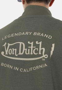 Sweat-shirt vert olive arborant le logo "Von Dutch" en beige clair, avec le texte "MARQUE LÉGENDAIRE" et "NÉ EN CALIFORNIE." Le texte est courbé autour du logo.