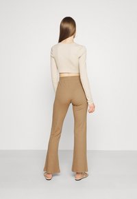 Beige kortärmad topp med långa ärmar, i kombination med bruna utsvängda byxor gjorda av ribbat tyg. Modellen står med ryggen mot betraktaren, framför en enfärgad bakgrund.