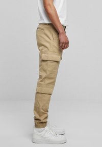 Khaki Cargo-Shorts mit mehreren Seitentaschen, schmal zulaufendem Knöchel und elastischen Bündchen; kombiniert mit weißen Turnschuhen und einem schlichten weißen Hemd.