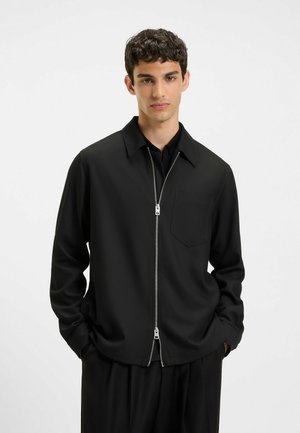 Black zip-up jacket with a pointed collar, long sleeves, a chest pocket, and smooth fabric texture. Simple, minimalist design.