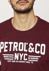 Bordeauxrotes Langarmshirt mit fettgedrucktem weißen Text: "PETROL&CO NYC NO BARRIERS." Weicher Stoff mit rundem Ausschnitt und lässiger Passform.