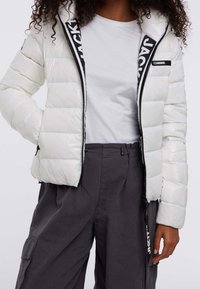 Giacca a vento bianca con design trapuntato, zip nera e dettagli neri, con cappuccio e logo ricamato sulla fodera interna.