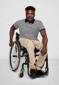 Moški na invalidskem vozičku, oblečen v polo majico z mornarsko modro in belo črtami ter kaki hlače, sedi z rokami na kolesih, vidni so črni čevlji.