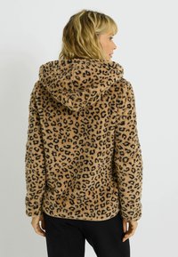 Sweat à capuche en polaire imprimé léopard couleur beige avec des taches noires. Texture douce, manches longues et grande capuche. Présente un ourlet arrondi et une coupe décontractée.