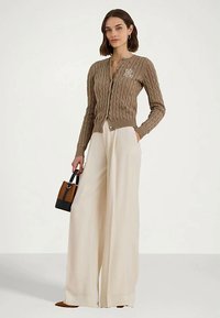 Γυναίκα με καφέ πλεκτό cardigan και κρεμ παντελόνι σε φαρδιά γραμμή, κρατώντας μια μικρή καφέ και μαύρη τσάντα.