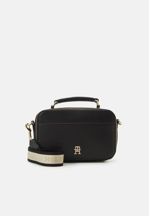 Sac bandoulière rectangulaire noir avec poignée sur le dessus, fermeture éclair dorée, logo doré et sangle beige épaisse avec bords noirs et lettres discrètes.