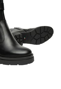 Stivali neri in pelle con suole di gomma spesse, uno stivale in posizione verticale e l'altro che mostra il motivo della suola e una piccola bandiera italiana.