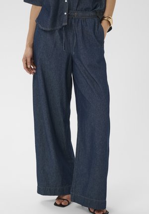 Žena má na sebe voľné široké nohavice z tmavomodrého denimu s elastickým pásom a sťahovacou šnúrkou, spolu s čiernymi otvorenými sandálmi a zlatým náramkom.