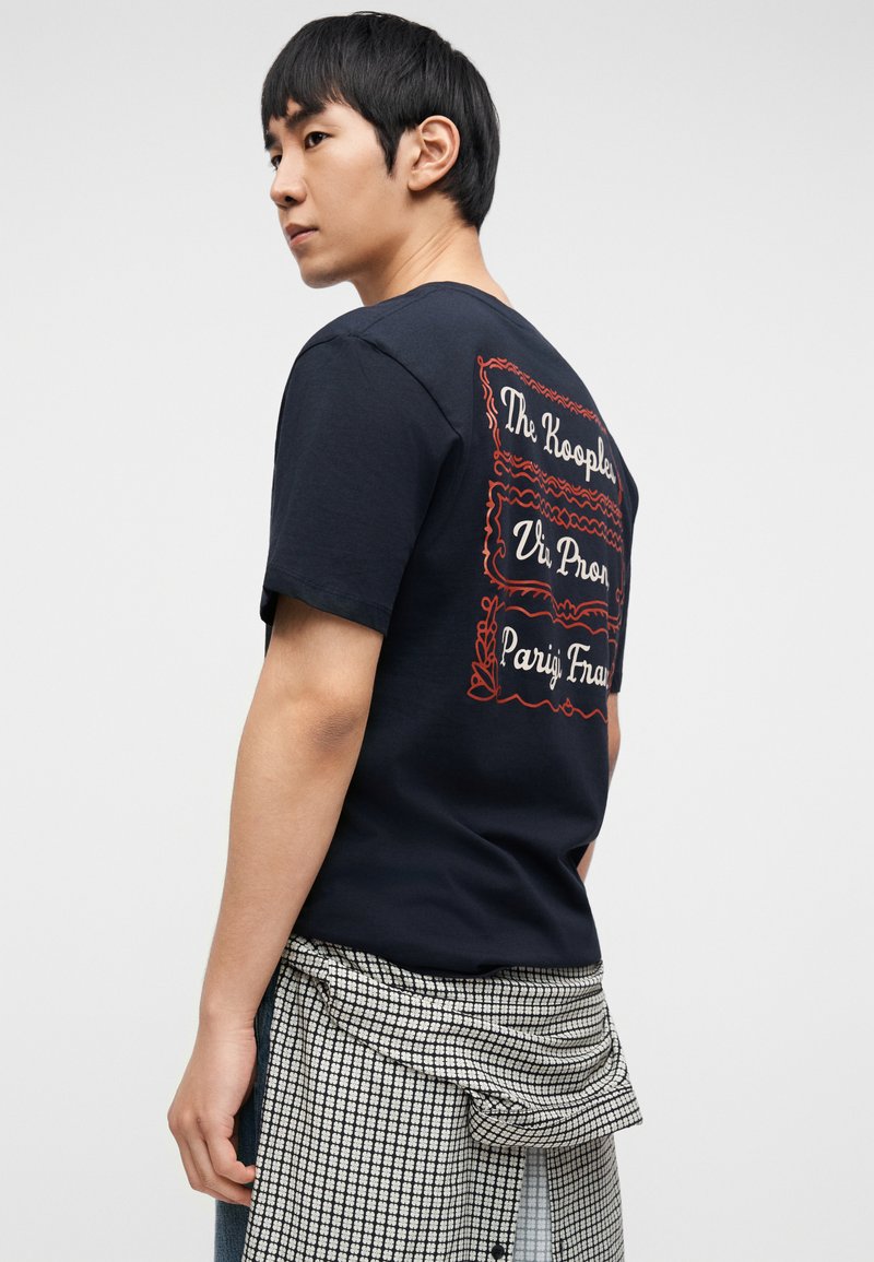 Navy cotton t-shirt featuring red graphic text on the back, with a relaxed fit. Layered with a checkered shirt tied at the waist.