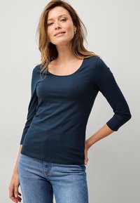 Marineblaues Langarmoberteil mit U-Boot-Ausschnitt, aus weichem Stoff gefertigt, hat eine taillierte Passform und einen leichten Stretch, kombiniert mit hellen Jeans.