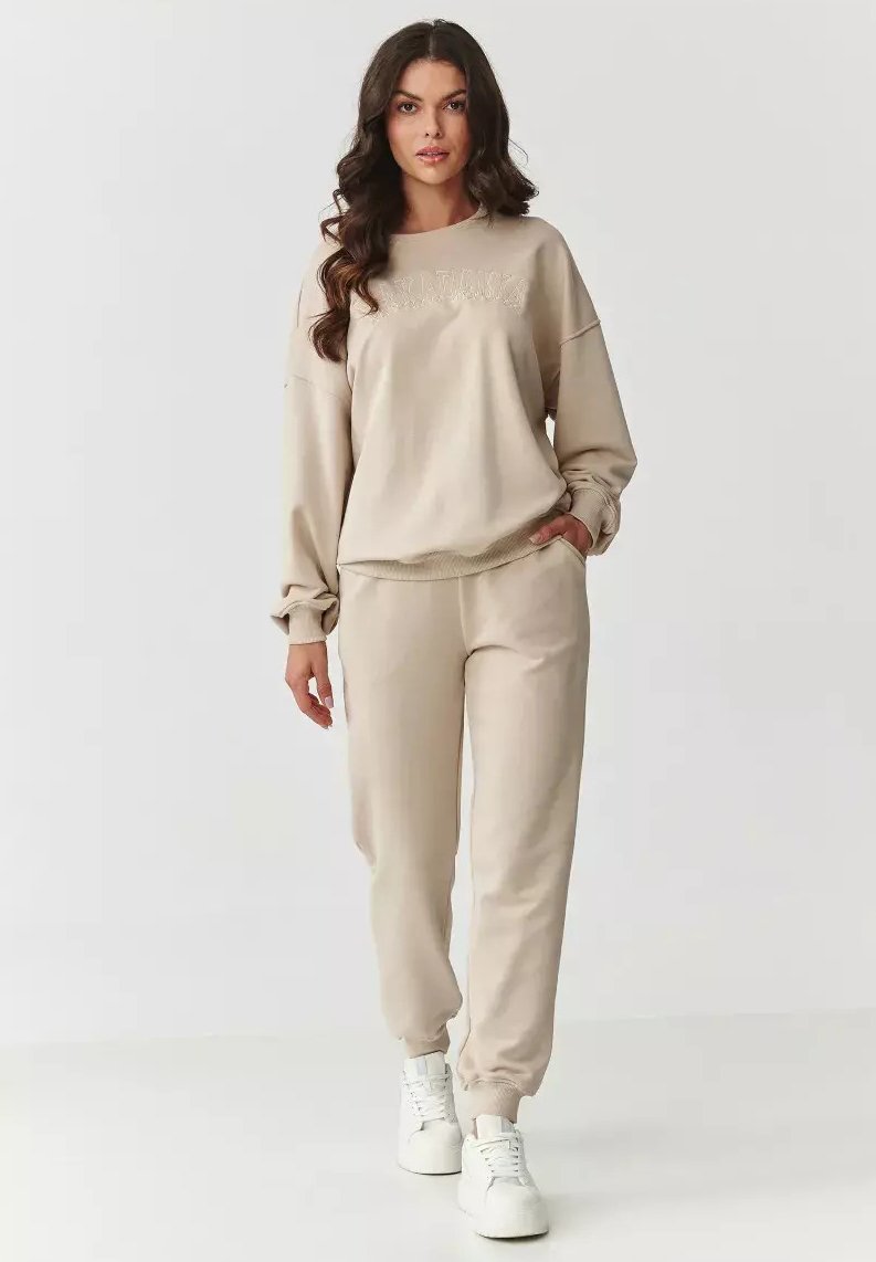 Beżowa bluza i pasujące spodnie dresowe wykonane z miękkiego materiału. Bluza ma wyszyty tekst; spodnie mają boczne kieszenie i ściągacze na rękawach.