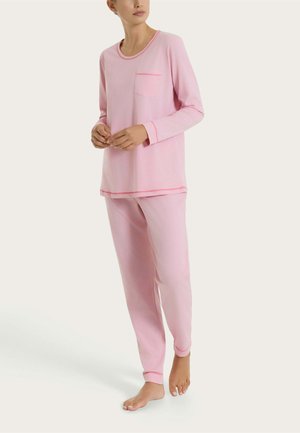 Donna che indossa una maglia a maniche lunghe rosa chiaro e pantaloni abbinati con rifiniture rosa, in piedi a piedi nudi su uno sfondo neutro.