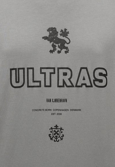 Sivá tričko s čiernou grafikou leva, veľkým tučným nápisom "ULTRAS" a ďalším textom a emblémom v menšom písme v dolnej časti.