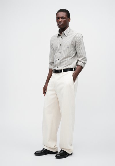 Striped skjorte med knapper i nyanser av hvit og grå, kombinert med løstsittende kremfargede bukser og svarte sko. Svart belte inkludert.