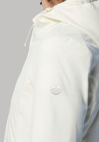 Giacca impermeabile bianca con cappuccio sagomato, texture liscia e logo circolare sulla manica sinistra. Nessuna chiusura visibile.