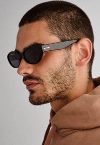 Óculos de sol pretos com lentes ovais e armações grossas, apresentando um detalhe em dourado nas laterais. O design é elegante e moderno.