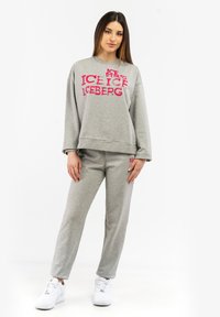 Grauer Sweatshirt und Hose aus weichem Material. Das Sweatshirt zeigt ein auffälliges pinkes Design mit der Aufschrift "ICEBERG". Das Model trägt weiße Sneaker.