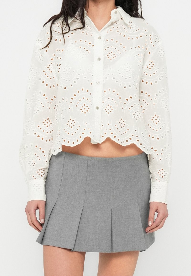 Femme portant une chemise blanche boutonnée en dentelle œillet avec un ourlet festonné et une mini-jupe plissée grise.