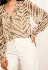 Blusa beige con motivo a zigzag marrone, scollatura arricciata e dettaglio con fiocco. Maniche lunghe con polsini arricciati. Abbinata a pantaloni bianchi.