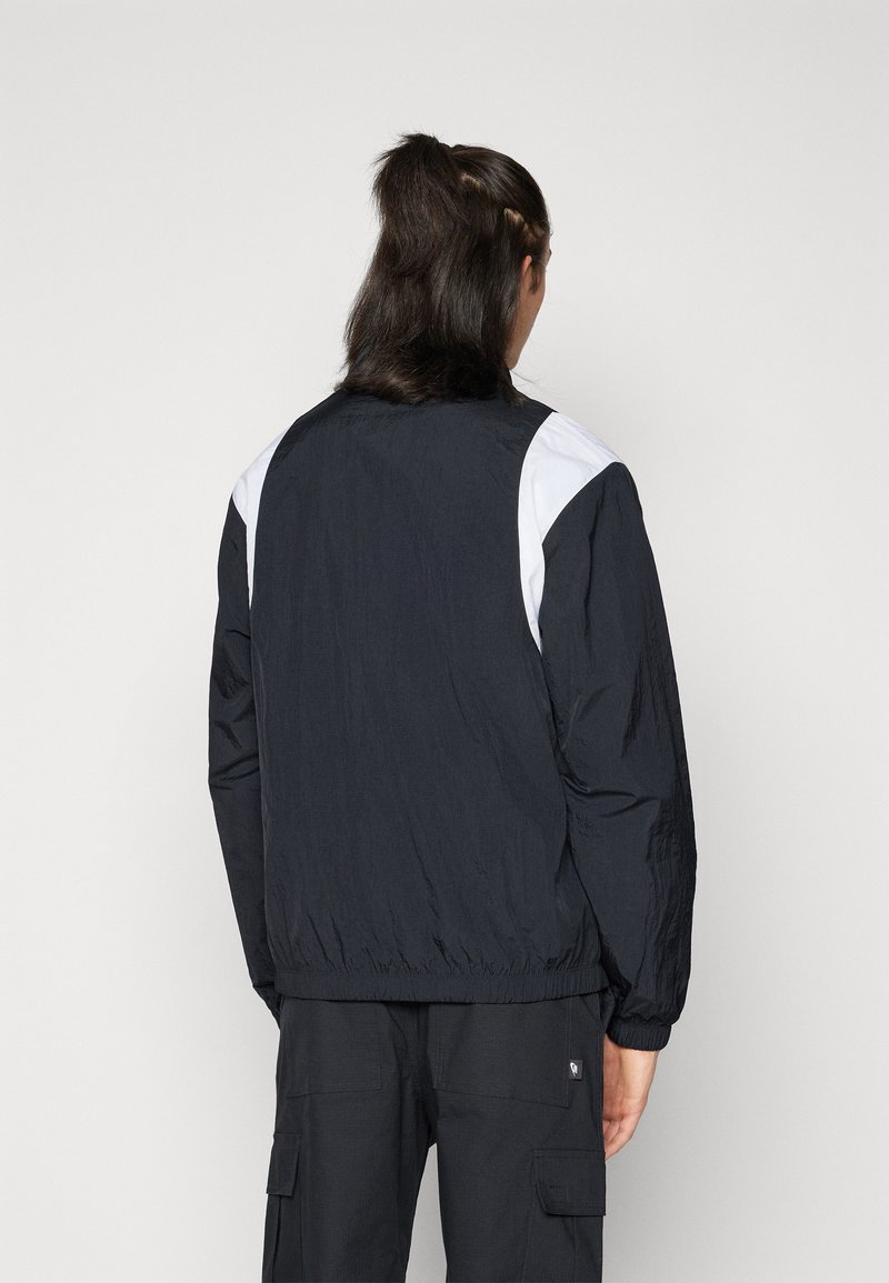 Veste légère noire avec des accents blancs sur les épaules, taille élastique et poignets côtelés. Tissu texturé avec une coupe décontractée.
