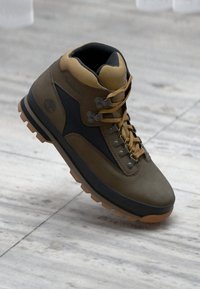 Túrabakancs barna velúrból készült felsőrésszel, fekete panelek, fém fűzőlyukak és gumitalp jellemzi. Fűzőkkel és párnázott nyakkal rendelkezik a támogatás érdekében.