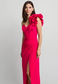 Fuksjowa sukienka na jedno ramię ozdobiona dużymi falbaniastymi akcentami. Prosta sylwetka z rozcięciem do uda i gładką teksturą.