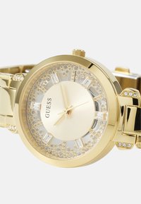 Zlatý kovový hodinky s okrúhlym ciferníkom, kryštálovými akcentmi a štruktúrovaným dialom. Má leštený kovový remienok a jednoduchú korunku na nastavovanie času.