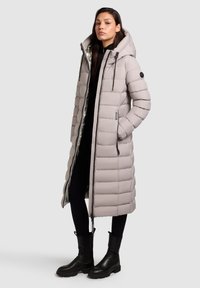 Gaiši pelēks pūkainais mētelis ar kapuci, ar garu dizainu, quilted faktūru un melniem rāvējslēdzēja elementiem. Valkāts virs melnām biksēm un masīvām apaviem.