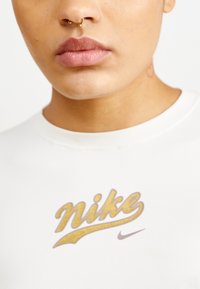 Balta medvilnės marškinėliai su apvalia apykakle, kuriuose matomas auksinis blizgus "Nike" logotipas ir mažas "Nike" swoosh detalė, arti žvilgsniu matoma apykaklė ir krūtinė.