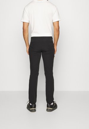 Mann trägt schwarze Slim-Fit-Hose, weißes T-Shirt, weiße Socken und schwarze Sneakers, steht mit dem Rücken zur Kamera auf grauem Boden und weißem Hintergrund.