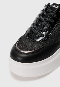 Svart sneakers med en teksturert, glitrende overdel; blanke patenterte skinnaksenter; tykk hvit såle; runde lisser og dekorative nagler.