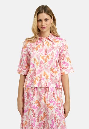 Frau, die ein passendes Shirt- und Rockset mit Tigerprint in Pink, Orange und Rot trägt, vor einem einfarbigen hellen Hintergrund stehend.