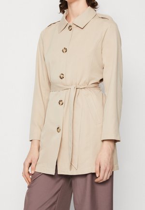 Veste beige ceinturée à manches longues avec de grands boutons en écaille de tortue portée sur un pantalon mauve à jambes larges.