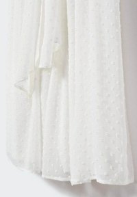 Vit genomskinlig tyg med texturerade prickar, lätt och flödande. Kanterna är lätt fransade, vilket ger en delikat känsla.