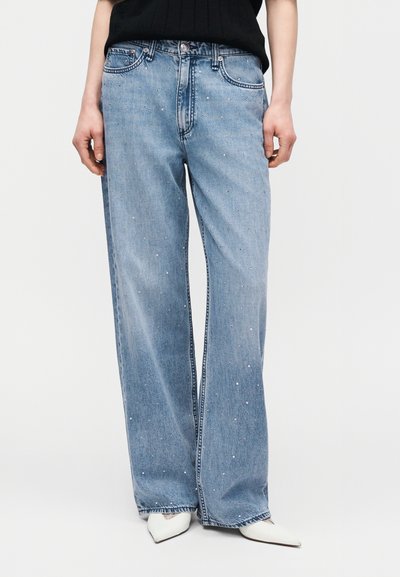 Φαρδιά τζιν παντελόνια ανοιχτού γαλάζιου με μικρές διάσπαρτες ασημένιες τρουκς, συνδυασμένα με μαύρο τοπ και λευκά παπούτσια με μυτερή μύτη.