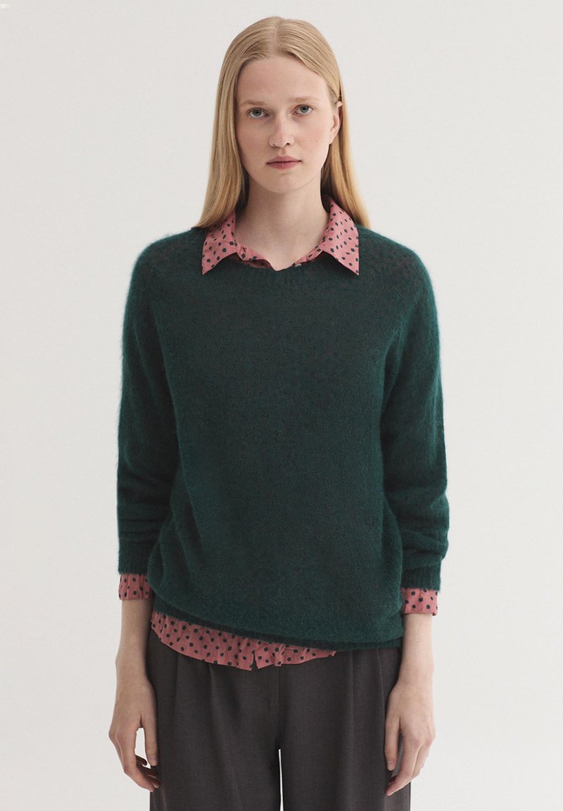 Jeune femme aux longs cheveux blonds portant un pull vert foncé sur une chemise rose à pois et un pantalon sombre, debout devant un fond uni.