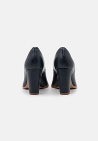 Une paire de chaussures à talons hauts en cuir noir avec des talons carrés vus de l'arrière sur un fond blanc.