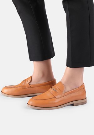 Fødder iført brune læderloafers kombineret med sorte cropped bukser på en hvid baggrund.