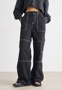 Persoon in zwarte wijde cargo-broek met witte stiksels en zwarte slippers, handen in de zakken, staand op een lichte vloer.