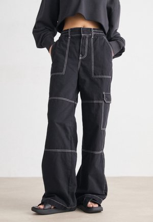 Person iført sorte vide cargo bukser med hvid syning og sorte slidesandaler, hænderne gemt i lommerne, stående på lys gulv.