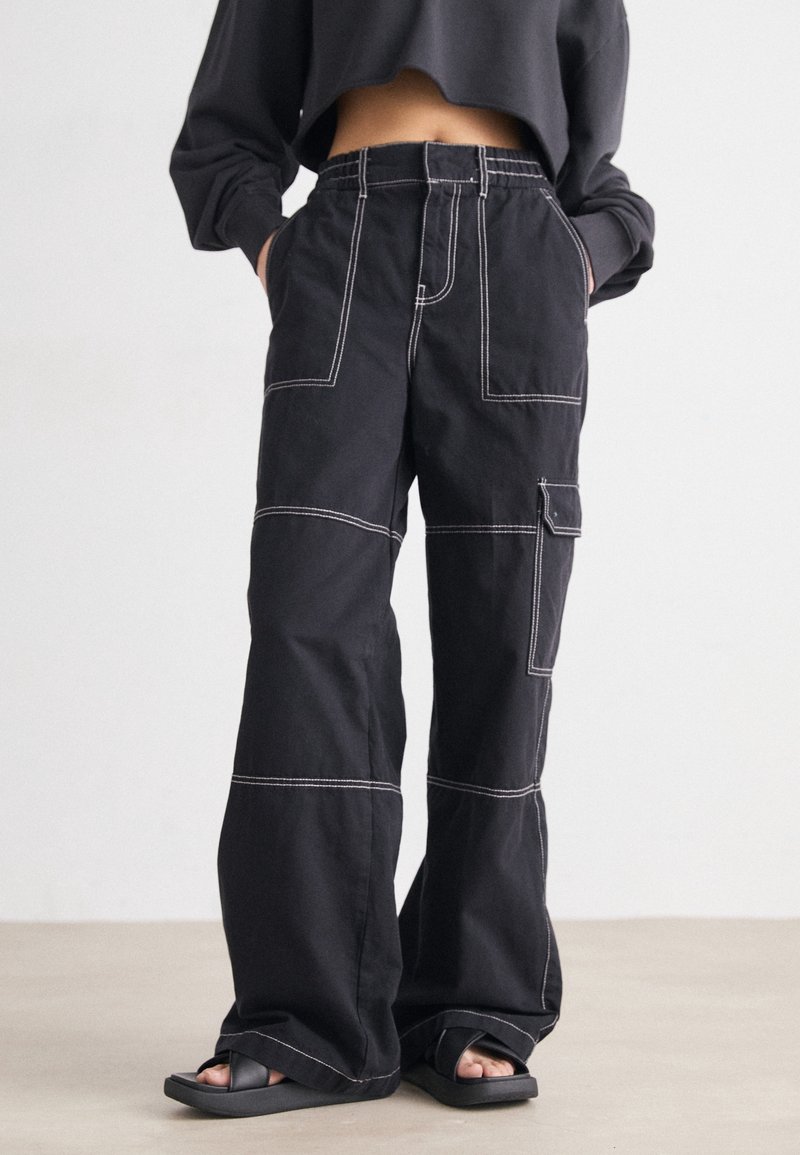 Persoon in zwarte wijde cargo-broek met witte stiksels en zwarte slippers, handen in de zakken, staand op een lichte vloer.